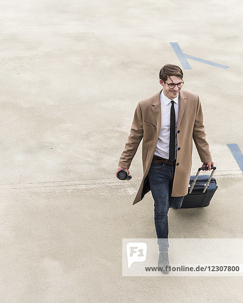 Smiling businessman with rolling suitcase walking at parking garage