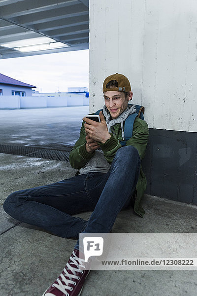 Lächelnder Mann sitzt auf dem Boden und schaut auf das Smartphone im Parkhaus.