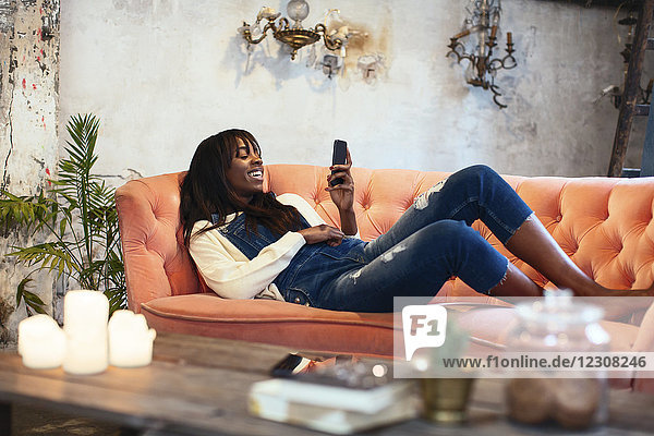 Lachende Frau zu Hause auf der Couch liegend mit dem Handy