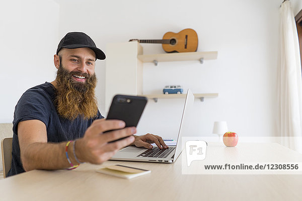 Portrait of bearded young man sitting at desk with laptop looking at cell phone