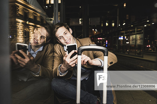 Lächelnder Geschäftsmann mit Handy an der Straßenbahnstation bei Nacht