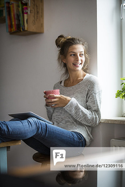 Lächelnde Frau auf Hocker sitzend mit Tasse und Tablette