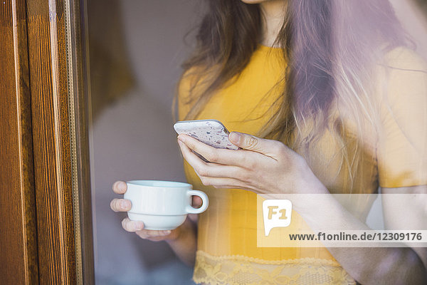 Junge Frau mit Kaffeetasse und Handy hinter Glas  Nahaufnahme