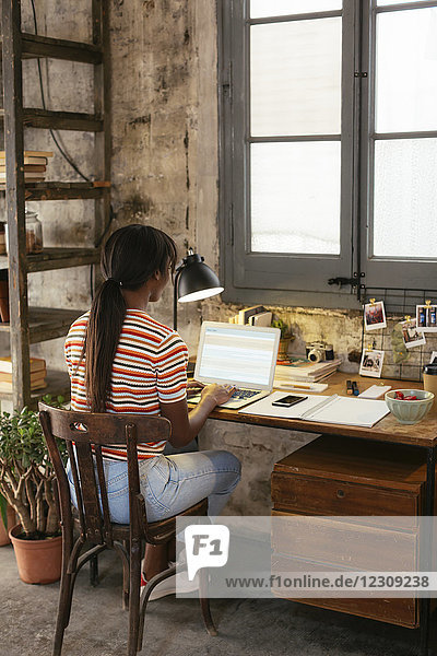 Rückansicht einer jungen Frau  die am Schreibtisch in einem Loft sitzt und am Laptop arbeitet.