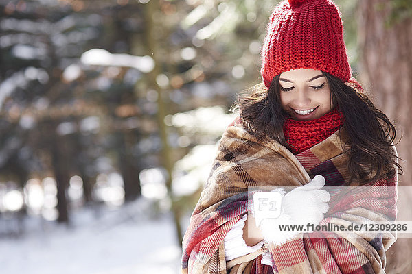 Lächelnde junge Frau in Decke gewickelt im Winterwald