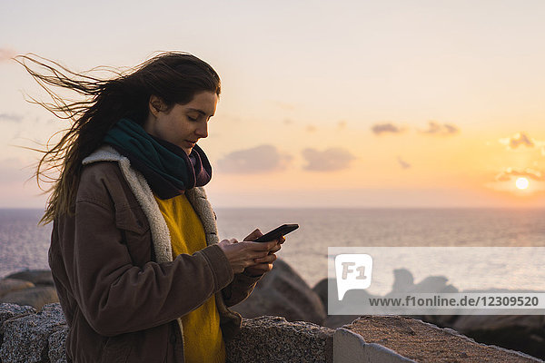Italien  Sardinien  Frau mit Handy an der Küste bei Sonnenuntergang