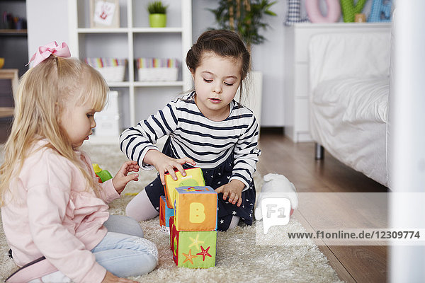 Zwei kleine Mädchen die zu Hause mit Spielzeugblöcken auf dem Teppich spielen.