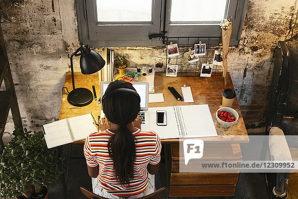 Back view of young woman sitting at desk in a loft working on laptop seen from above