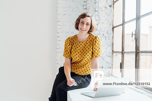 Porträt einer Geschäftsfrau  die mit einem Laptop auf der Fensterbank sitzt