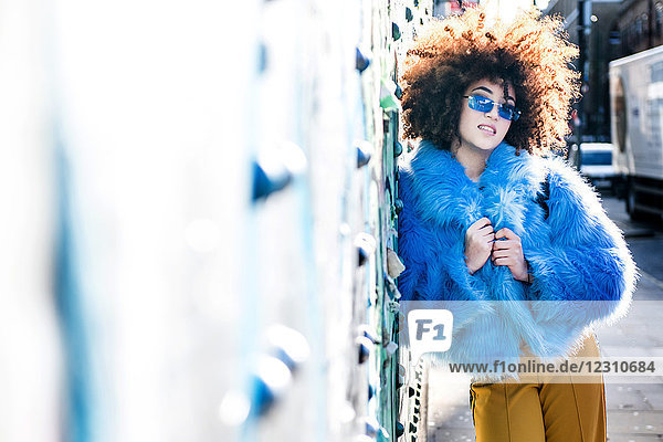 Portrait of woman with afro hair wearing fur coat  leaning against wall looking at camera