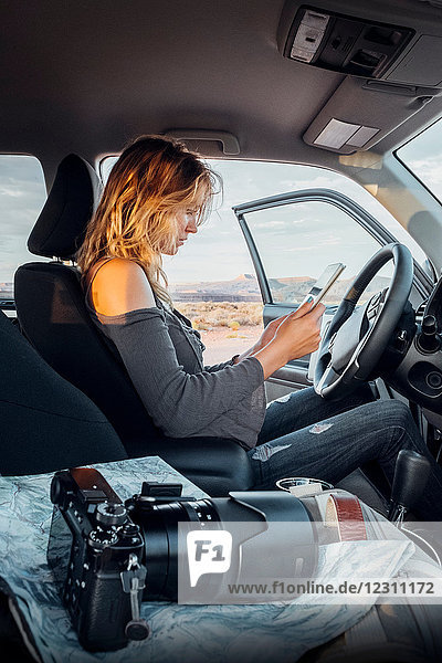 Junge Frau sitzt im Fahrzeug und schaut auf ein digitales Tablett  Spiegelreflexkamera auf dem Beifahrersitz  Mexican Hat  Utah  USA