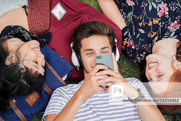 Young man lying on grass with friends  using smartphone and listening to headphones