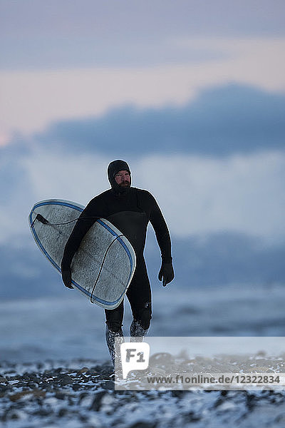 Mann mit Surfbrett auf dem Schnee am Strand von Homer im Winter  Süd-Zentral-Alaska  USA