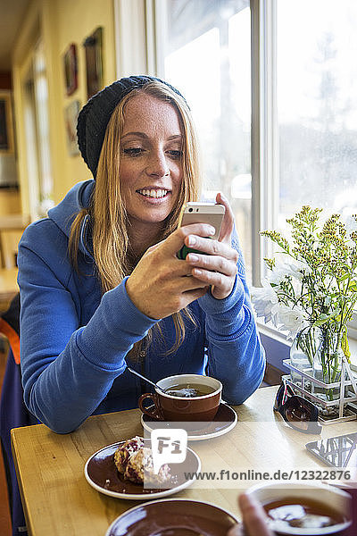 Junge Frau genießt eine Tasse Tee und Gebäck  während sie in einem Café eine SMS auf ihrem Smartphone schreibt  Süd-Zentral-Alaska; Anchorage  Alaska  Vereinigte Staaten von Amerika