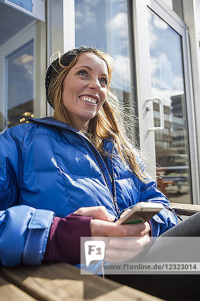 Junge Frau sitzt mit ihrem Smartphone vor einem Einkaufszentrum in der Innenstadt von Anchorage  Süd-Zentral-Alaska; Anchorage  Alaska  Vereinigte Staaten von Amerika