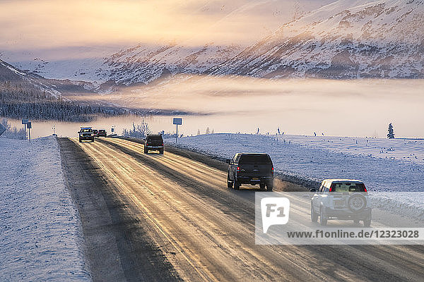 Ein nebliger Sonnenuntergang beleuchtet Autos  die den schneebedeckten Seward Highway hinunterfahren  die Kenai Mountains im Hintergrund  Kenai-Halbinsel  Süd-Zentral-Alaska im Winter; Alaska  Vereinigte Staaten von Amerika