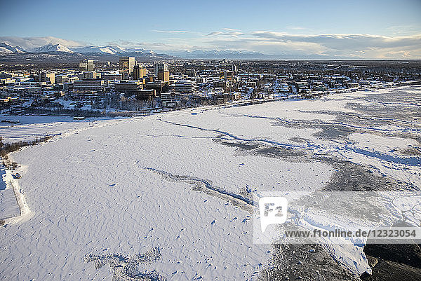 Luftaufnahme von Schnee  der das Meereis an den gefrorenen Ufern der Innenstadt von Anchorage bedeckt  die Chugach Mountains im Hintergrund hinter den Bürogebäuden und Hotels in der Innenstadt von Anchorage im Winter  Süd-Zentral-Alaska; Anchorage  Alaska  Vereinigte Staaten von Amerika