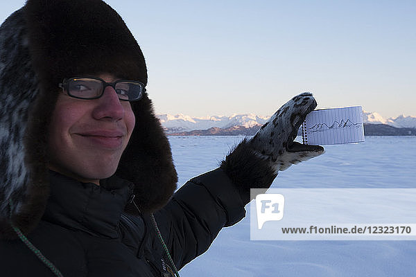 Young man showing off drawing of Kenai Mountains and holding notepad with the range in the background while standing in the snow at sunset  South-central Alaska; Alaska  United States of America