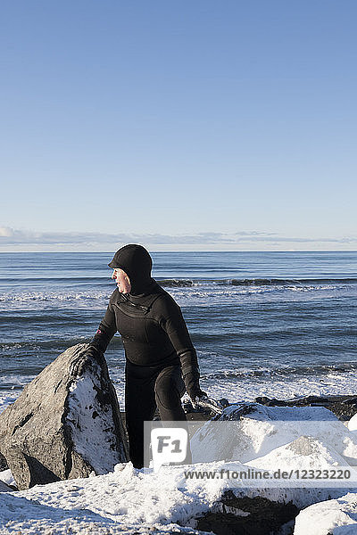 Female surfer in a wetsuit walking among the rocks and snow on the Homer Spit shoreline in winter  South-central Alaska; Homer Spit  Alaska  United States of America