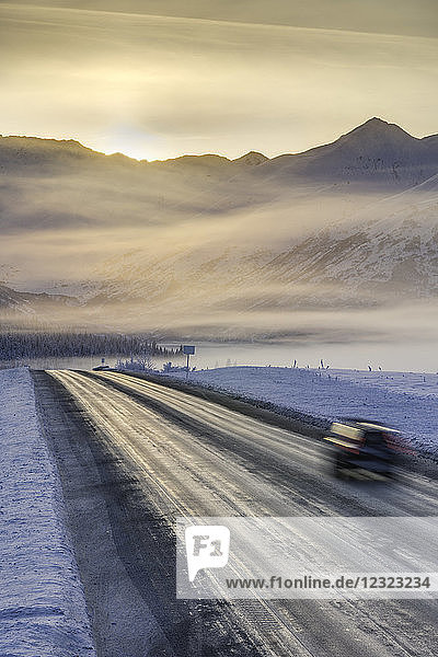 Ein Auto fährt eine Straße in einer schneebedeckten Landschaft hinunter  die Sonne geht über einem Gebirgskamm im Hintergrund auf  Nebel verdunkelt die Silhouette im Winter  Turnagain Pass  Kenai-Halbinsel  Süd-Zentral-Alaska; Alaska  Vereinigte Staaten von Amerika