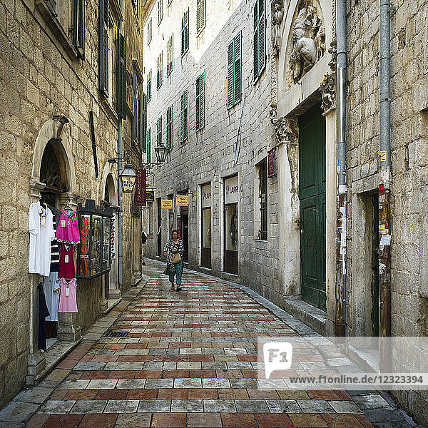 A woman walks down a narrow street between buildings with clothing on display outside a shop; Kotor  Montenegro