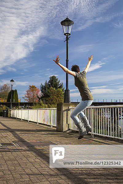 Eine junge Frau springt mit ausgestreckten Armen auf einem Pfad in einem Park in die Luft; New Westminster  British Columbia  Kanada