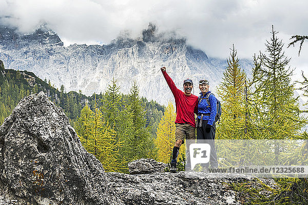 Male and female hikers standing on rock with autumn coloured trees and cloud-covered mountain range in the background; Sesto  Bolzano  Italy