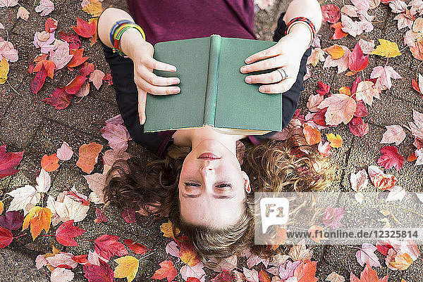 Eine junge Frau liegt auf dem Rücken auf dem Boden mit gefallenen Herbstblättern und liest ein Buch; New Westminster  British Columbia  Kanada