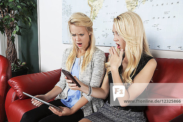 Zwei hübsche junge Geschäftsfrauen der Millennials sitzen an einem Arbeitsplatz und nutzen ihre Technologie mit schockierten Gesichtern  während sie etwas in den sozialen Medien lesen; Sherwood Park  Alberta  Kanada