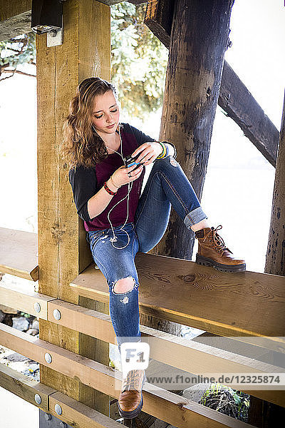 Eine junge Frau sitzt auf einem Holzgeländer unter einer Brücke am Wasser und hört Musik; New Westminster  British Columbia  Kanada