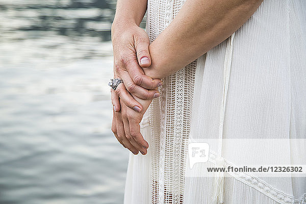 A woman's hands held together in front of a white dress while standing in water; British Columbia  Canada