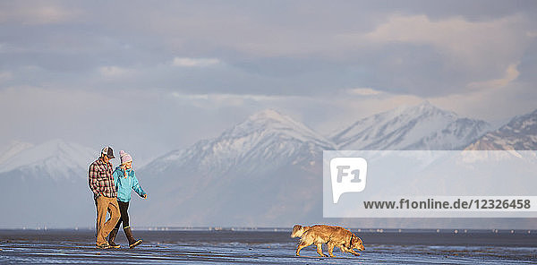 Ein junges Paar  das mit seinem Hund am Strand spazieren geht und Händchen hält  mit Blick auf eine Bergkette in der Ferne; Anchorage  Alaska  Vereinigte Staaten von Amerika