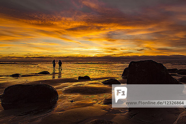 Silhouette of two people standing on Wreck Beach with an orange glow at sunset; Vancouver  British Columbia  Canada