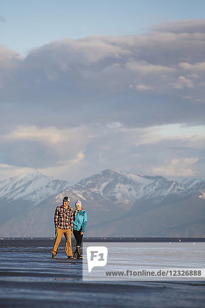 Ein junges Paar  das an einem Strand spazieren geht und sich an den Händen hält  mit einer Bergkette in der Ferne; Anchorage  Alaska  Vereinigte Staaten von Amerika