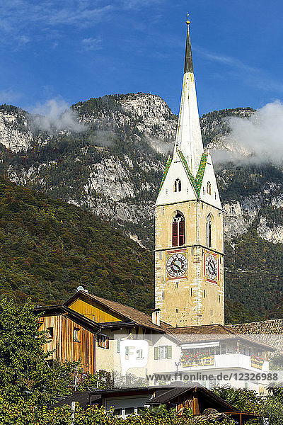 Uhrenturm einer Steinkirche in einem Alpendorf vor einem Berg mit blauem Himmel; Kalterer  Bozen  Italien