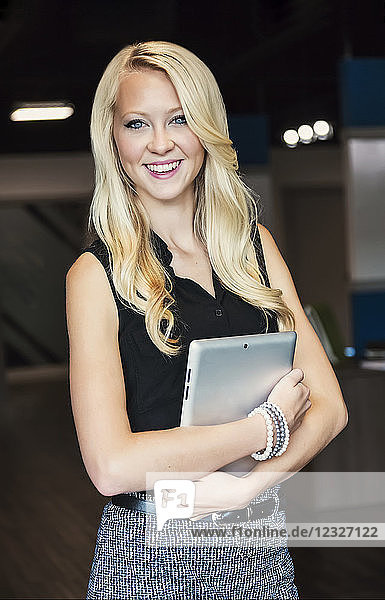 Business portrait of a beautiful young millennial businesswoman with long blond hair holding a tablet and posing for the camera in the workplace; Sherwood Park  Alberta  Canada