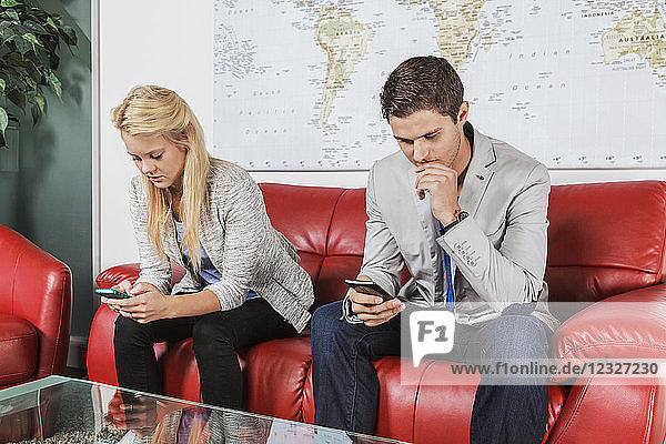 Zwei junge Geschäftsleute in der Lobby ihres Arbeitsplatzes  die in einer Pause ihre Smartphones benutzen; Sherwood Park  Alberta  Kanada