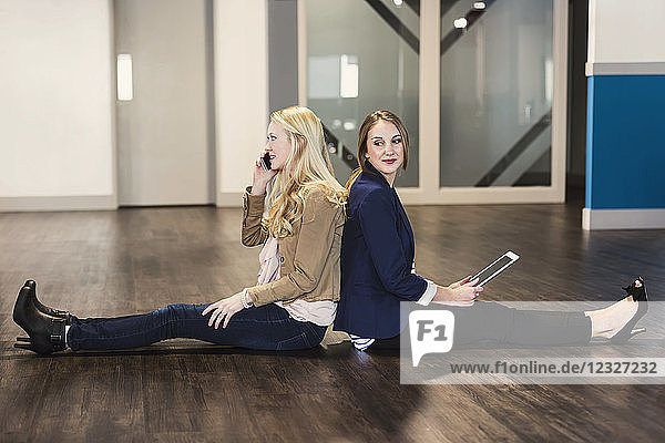 Zwei hübsche junge Frauen  die als Geschäftsleute der Jahrtausendwende Rücken an Rücken auf dem Boden ihres Arbeitsplatzes sitzen und Technologie nutzen; Sherwood Park  Alberta  Kanada
