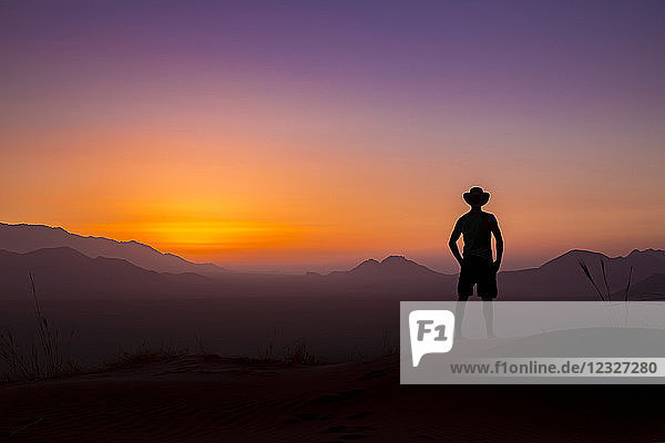 Silhouette eines Mannes mit Cowboyhut  der bei Sonnenuntergang auf die leuchtende Landschaft und den Himmel blickt; Sossusvlei  Hardap-Region  Namibia