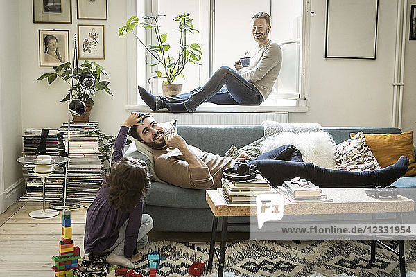 Girl playing with fathers while man sitting on window sill in living room