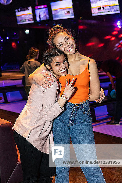 Portrait of smiling teenage girl showing peace sign while embracing friend at bowling alley