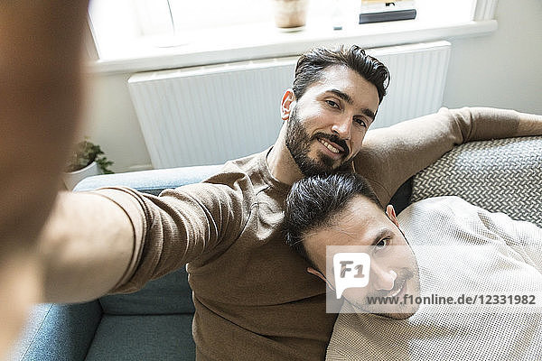 Smiling gay couple taking selfie while relaxing on sofa in living room at home