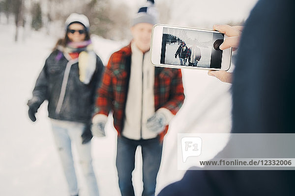 Woman photographing friends through smart phone on field during winter