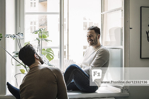 Happy gay man talking to boyfriend while sitting on windowsill at home