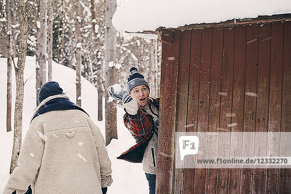 Man throwing snowball while standing with friend by log cabin during winter