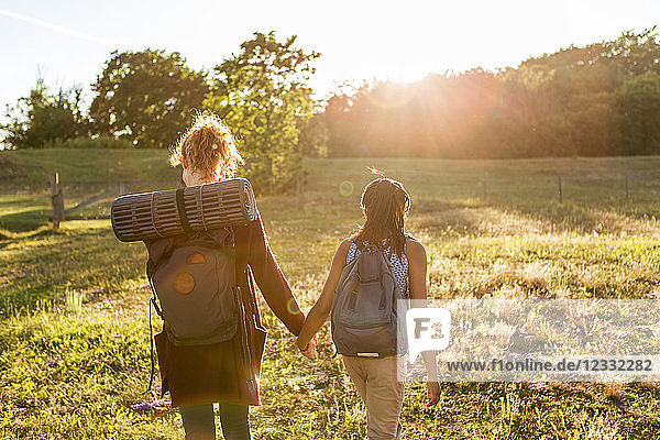 Rear view of girl and woman with backpacks holding hands while hiking on grass at public park during sunset