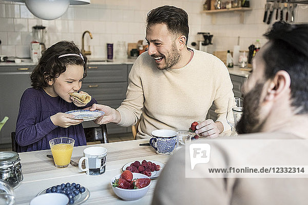 Laughing father holding plate while daughter eating pancake at table