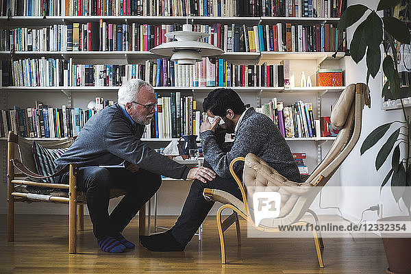 Senior therapist counseling young patient while sitting by bookshelf during therapy session