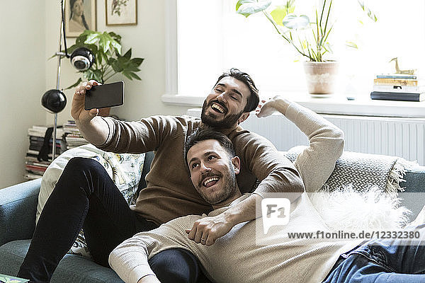 Happy gay couple taking selfie through mobile phone while relaxing on sofa at home