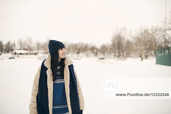 Smiling woman looking away while standing on snowy field during winter